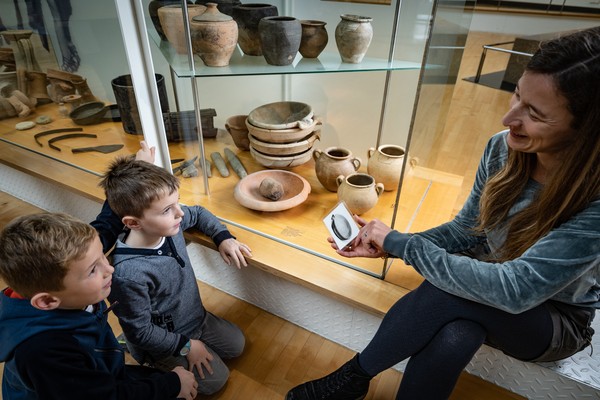 Foto: Mithilfe einer Suchkarte sind zwei Jungen gemeinsam mit einer Museumspädagogin zur Vitrine mit der Reibschüssel gelangt.